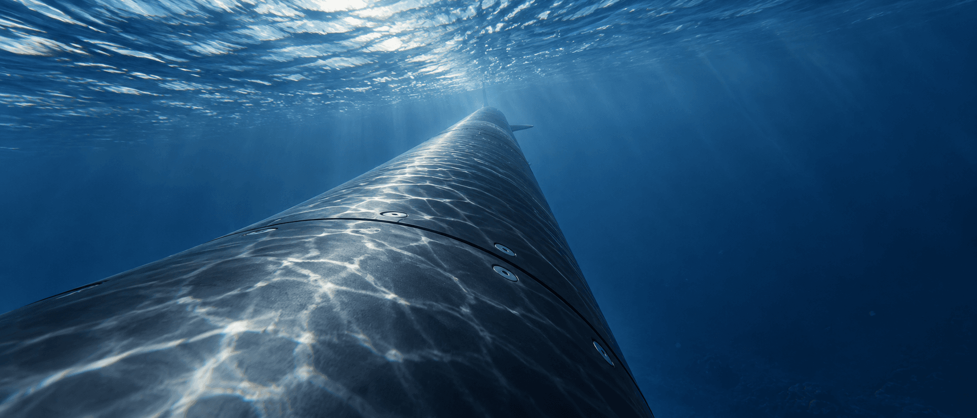 Illustrated close view along an underwater vehicle's hull, with reflected sunlight beneath the rippling sea surface.