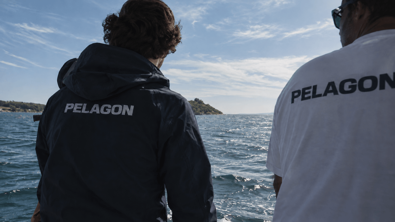 Two crew members seen from behind aboard a boat, wearing Pelagon clothing and looking out across the sea.